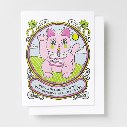 Risograph greeting card with an illustrated pink lucky cat holding a birthday cake in a decorative oval frame with clovers and horseshoes, text reads Hey Birthday Cutie You Deserve All the Luck on a pink banner