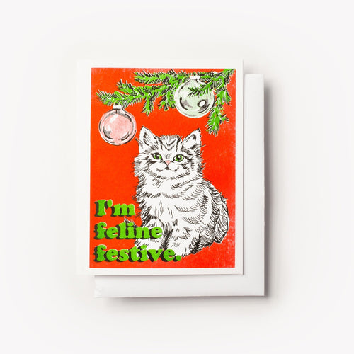 Risograph greeting card with an illustrated fluffy white cat sitting under pine tree branches with ornaments on a red background, text reads I'm Feline Festive in bold green lettering
